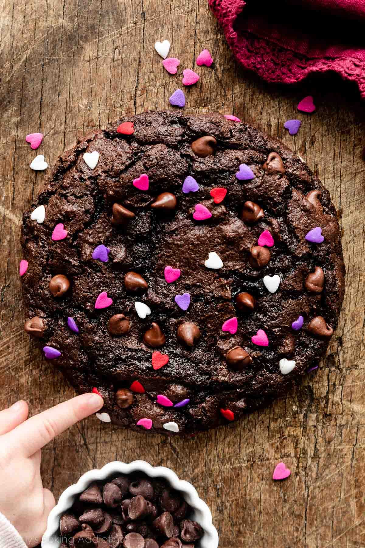 giant chocolate cookie with chocolate chips and Valentine's Day heart sprinkles on wooden cutting board with small hand pointing to it.