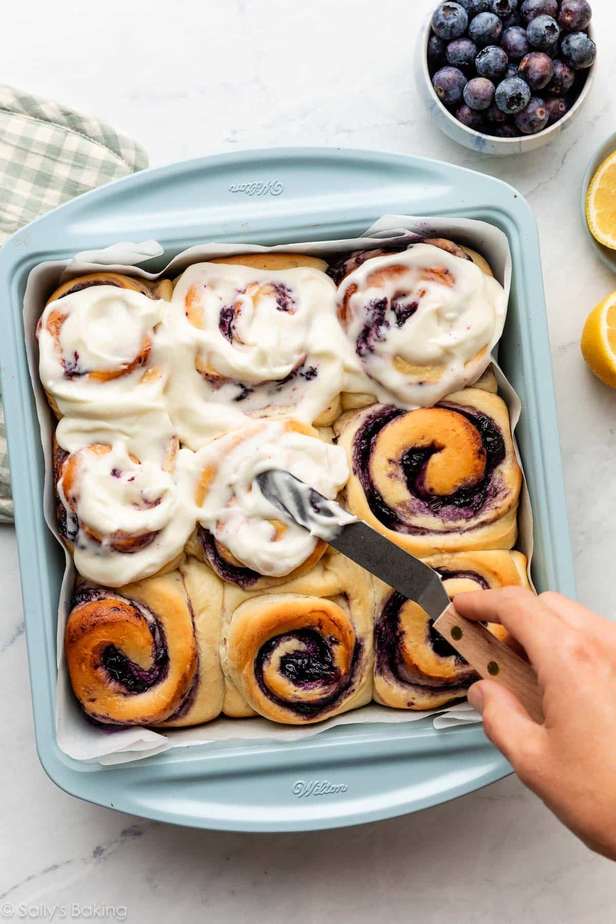 spreading frosting on blueberry rolls in blue pan.