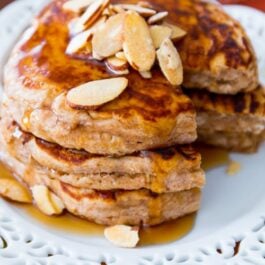 stack of whole wheat oatmeal pancakes topped with slivered almonds and maple syrup on a white plate