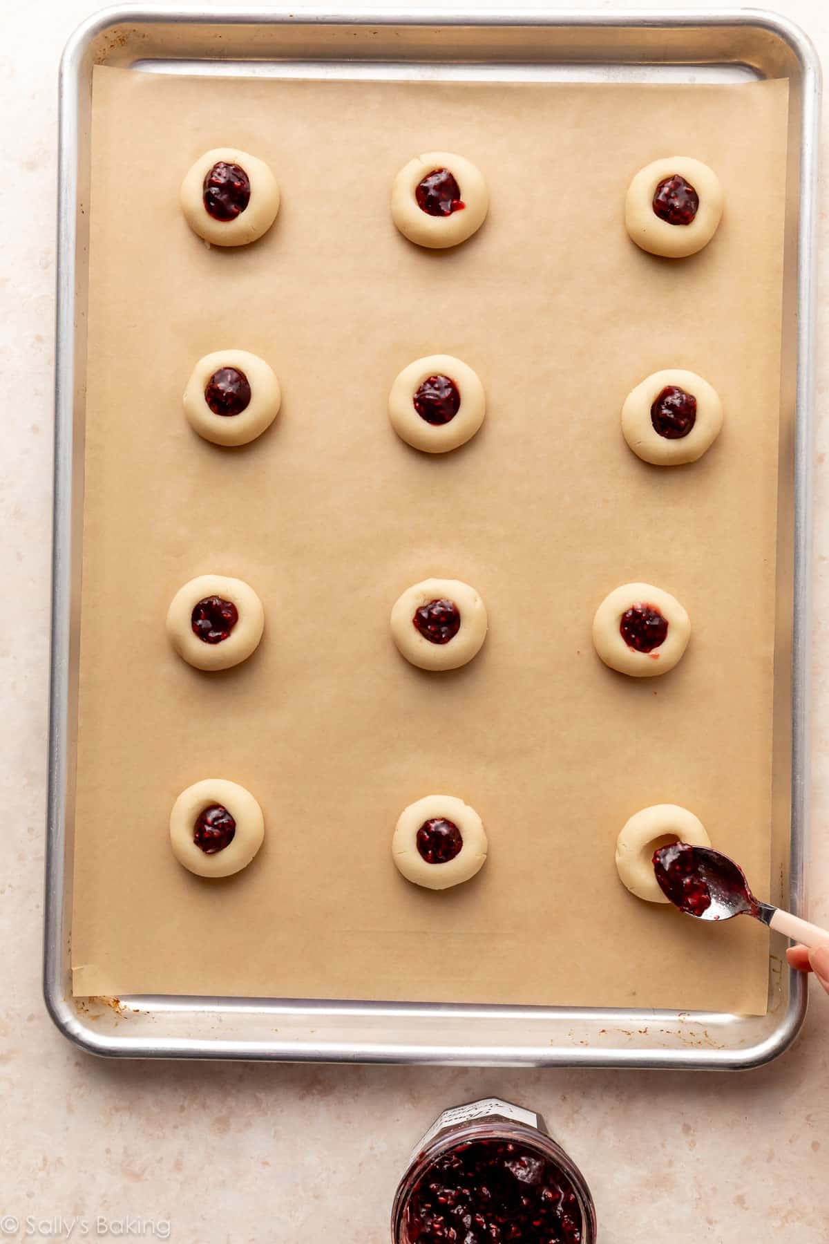 spooning raspberry jam into shaped thumbprint cookie dough.