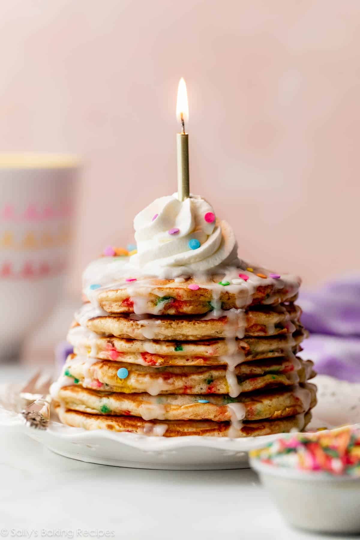 stack of birthday cake funfetti pancakes with icing dripping down the sides, whipped cream on top, and a lit gold candle on top.