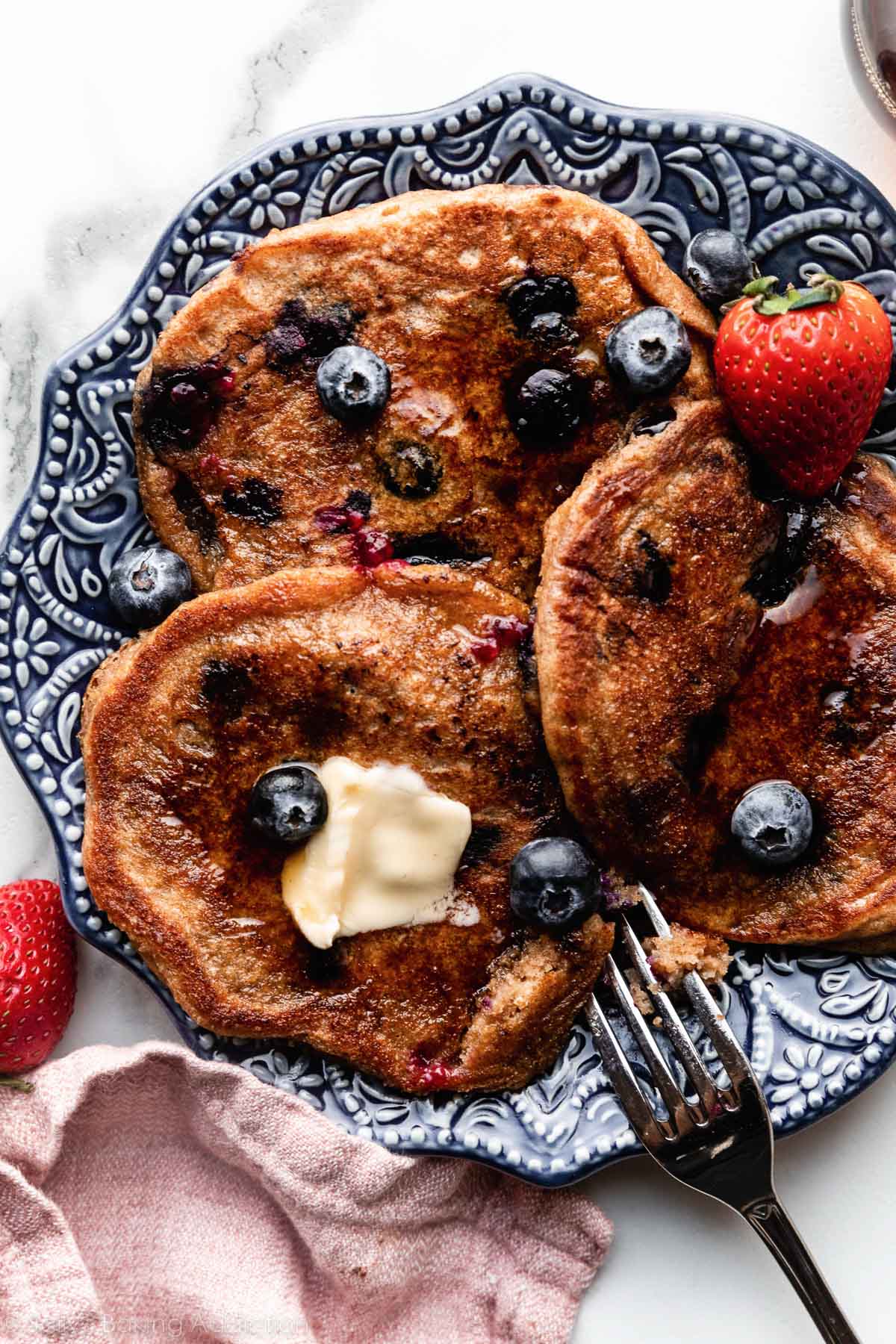 3 whole wheat blueberry pancakes on blue patterned plate with maple syrup on top.