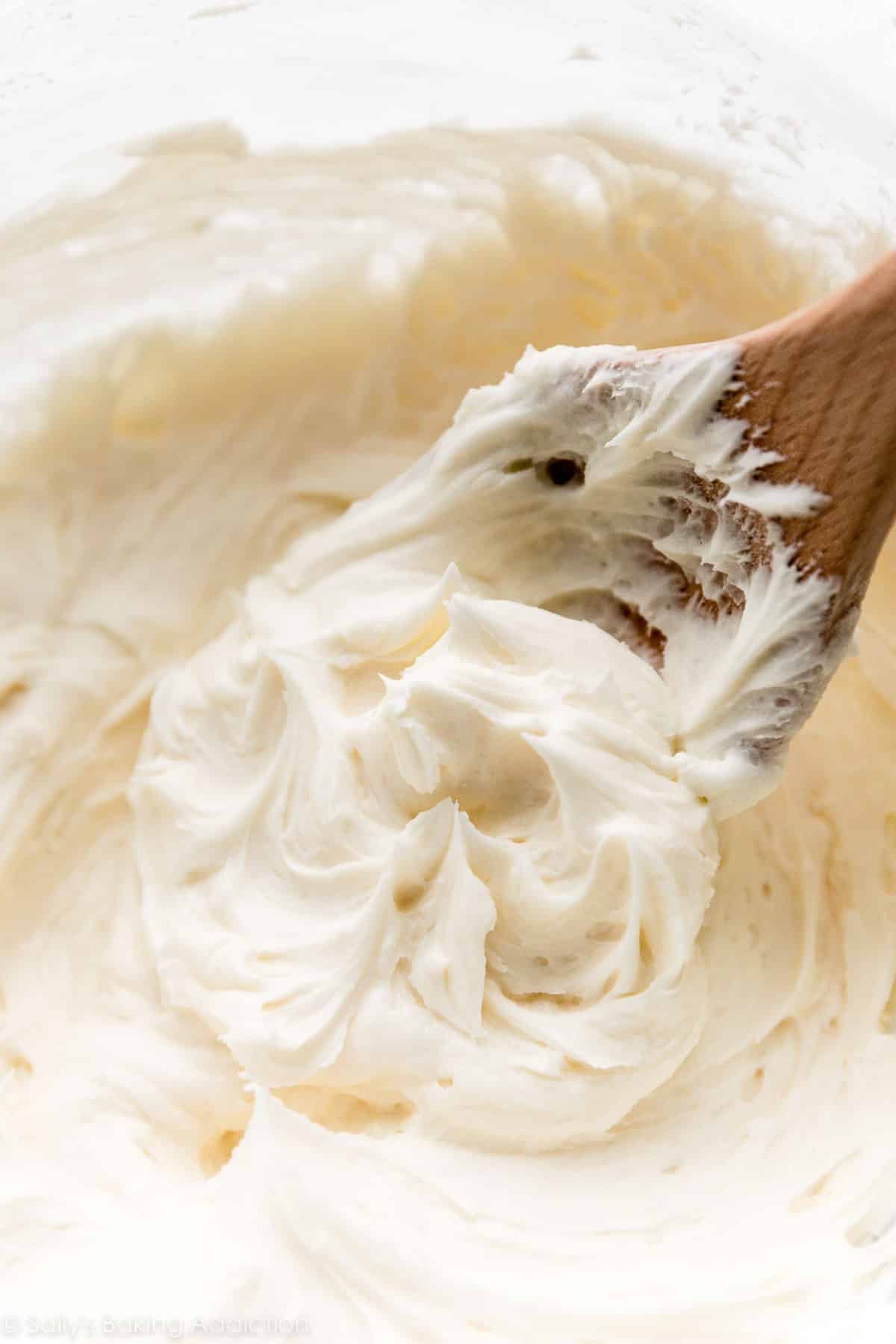 vanilla buttercream frosting being stirred by wooden spoon in big glass mixing bowl.