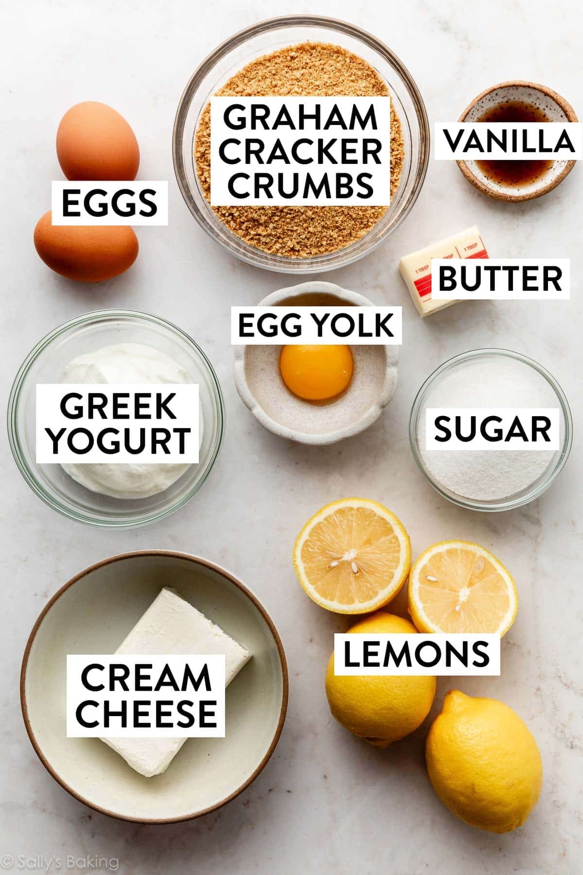 ingredients on marble surface including graham cracker crumbs, eggs, egg yolk, sugar, lemons, and cream cheese.
