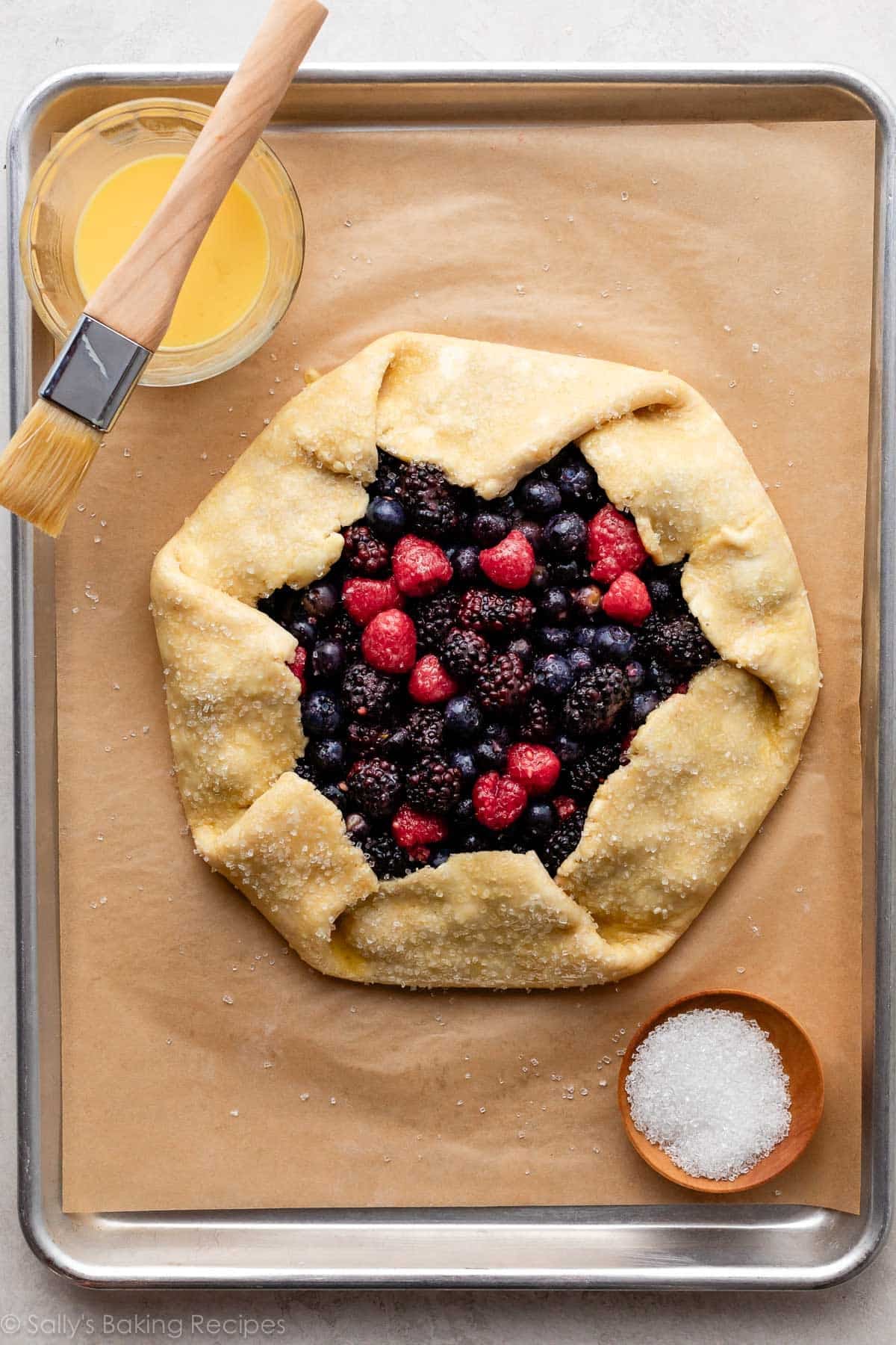 galette with berries on lined baking sheet with egg wash and pastry brush in top corner.