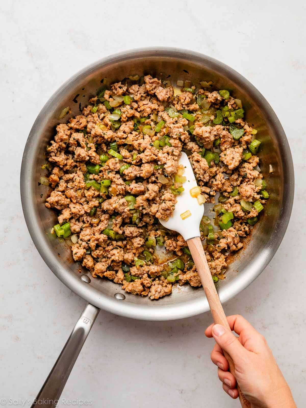 hand holding white spatula stirring cooked sausage and celery.