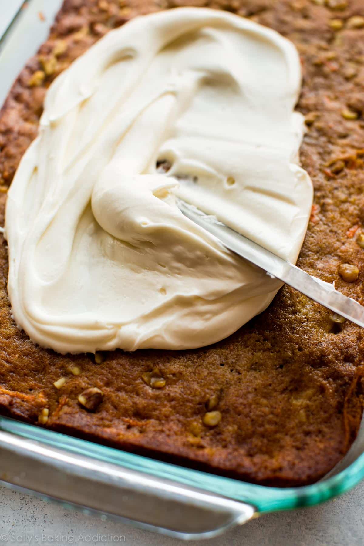 spreading cream cheese frosting onto pineapple carrot cake in a glass baking pan