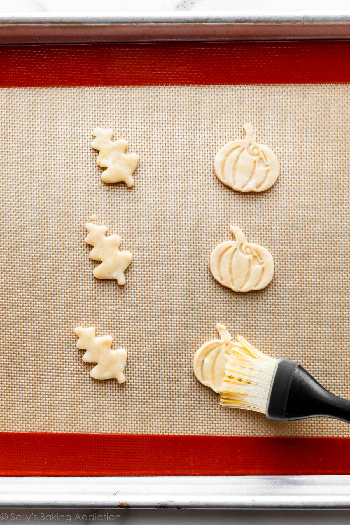 brushing egg wash on pie dough pumpkin shape with other pumpkin shapes and leaves on a silicone mat-lined baking sheet.