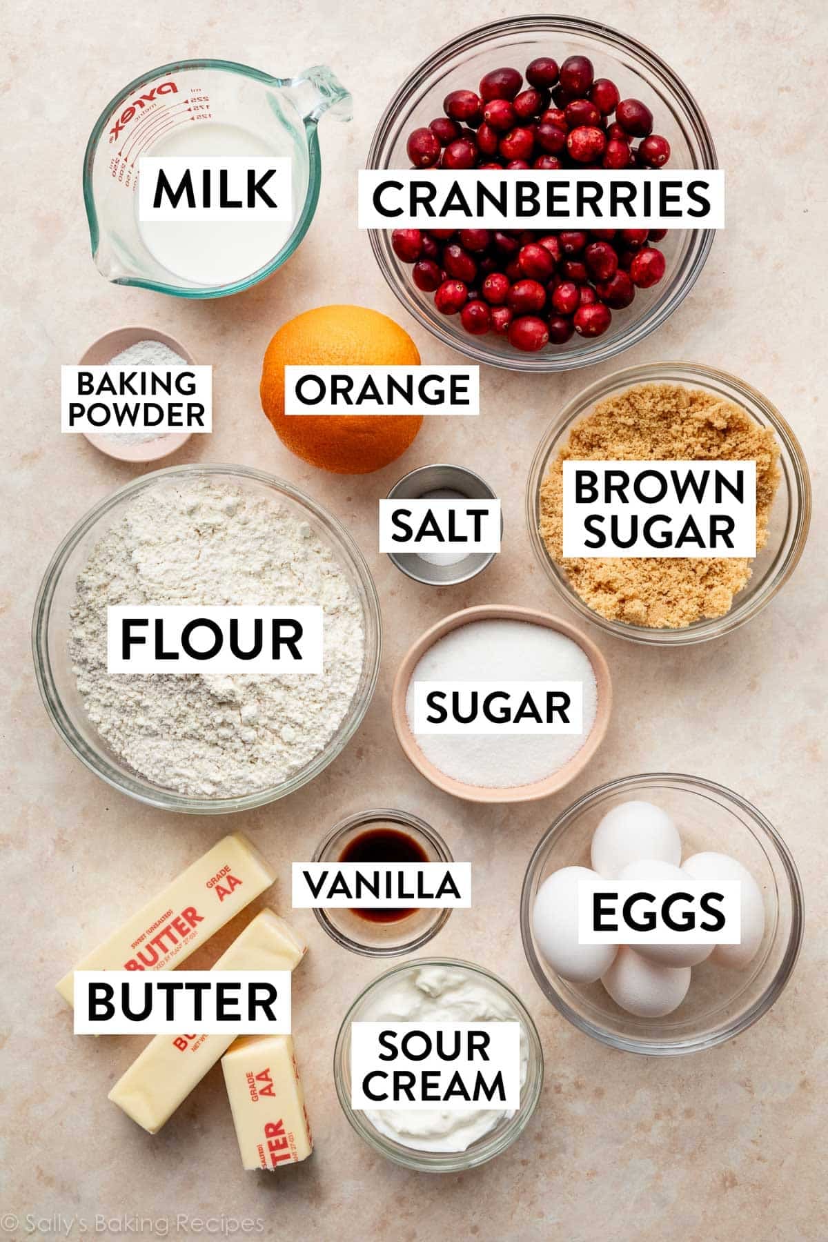 ingredients on beige surface including eggs, sugar, flour, brown sugar, butter, sour cream, vanilla, and orange.