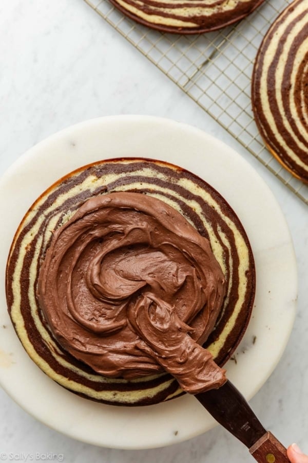 large icing spatula spreading chocolate frosting on zebra cake layer.