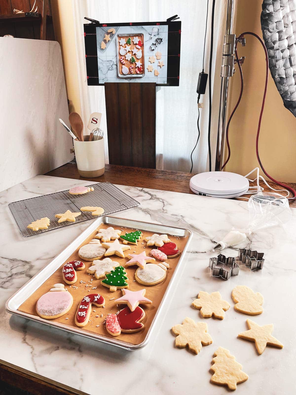 decorated Christmas sugar cookies on marble board with cookie cutters and monitor screen behind it.