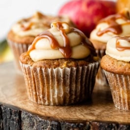 apple cupcakes topped with salted caramel frosting and caramel sauce sitting on wooden cake stand.