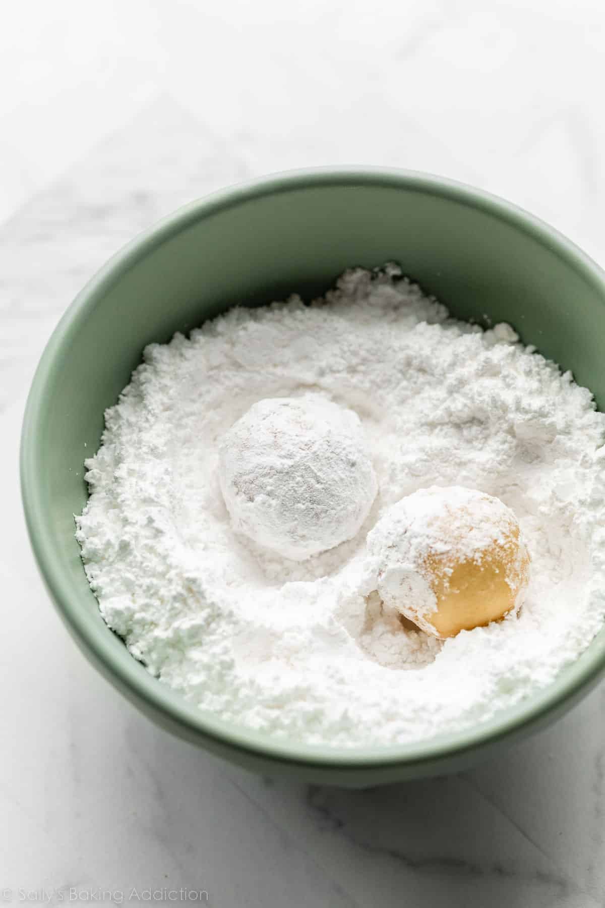 two dough balls rolled in green bowl of confectioners' sugar.
