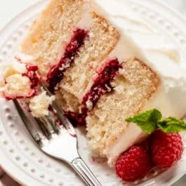 3 layer white vanilla cake slice on white plate with vanilla frosting and raspberry cake filling in the layers.