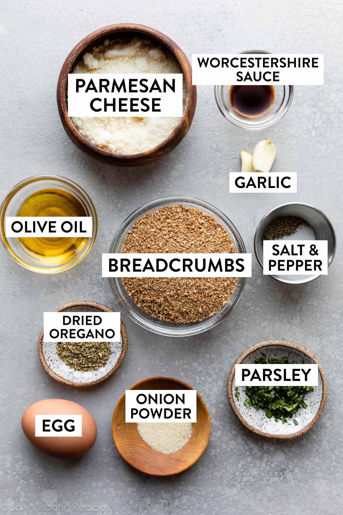 ingredients in bowls on gray backdrop including breadcrumbs, parmesan cheese, garlic, salt and pepper, herbs, egg, and olive oil.