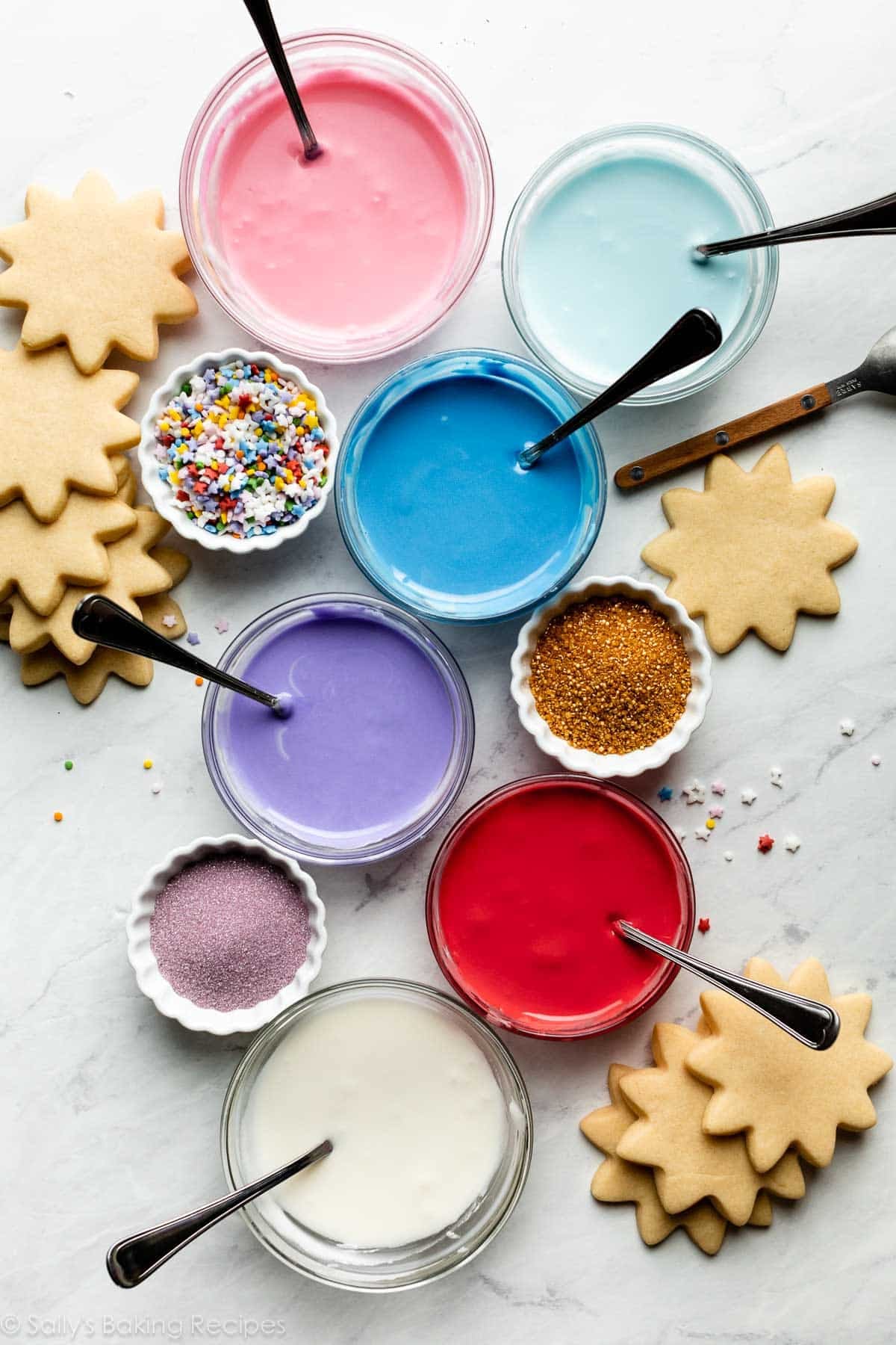 bowls of colored icing including pink, light blue, dark blue, purple, red, and white with bowls of sprinkles arranged around them.