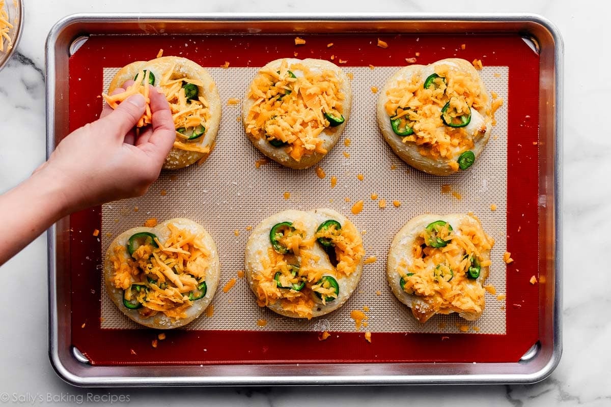 hand sprinkling cheese on top of jalapeño slices on par-baked pretzels.