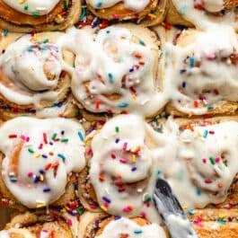 spreading cream cheese icing on sprinkle funfetti cake cinnamon rolls.