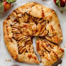 apple galette with caramel sauce on white parchment paper with apples and jar of caramel.