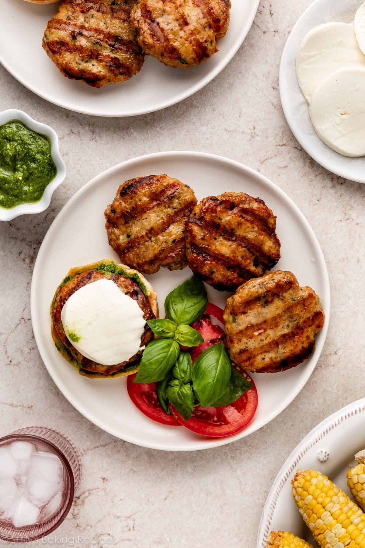 plate of burgers with grill marks with tomatoes and fresh basil, and a bowl of pesto next to it.