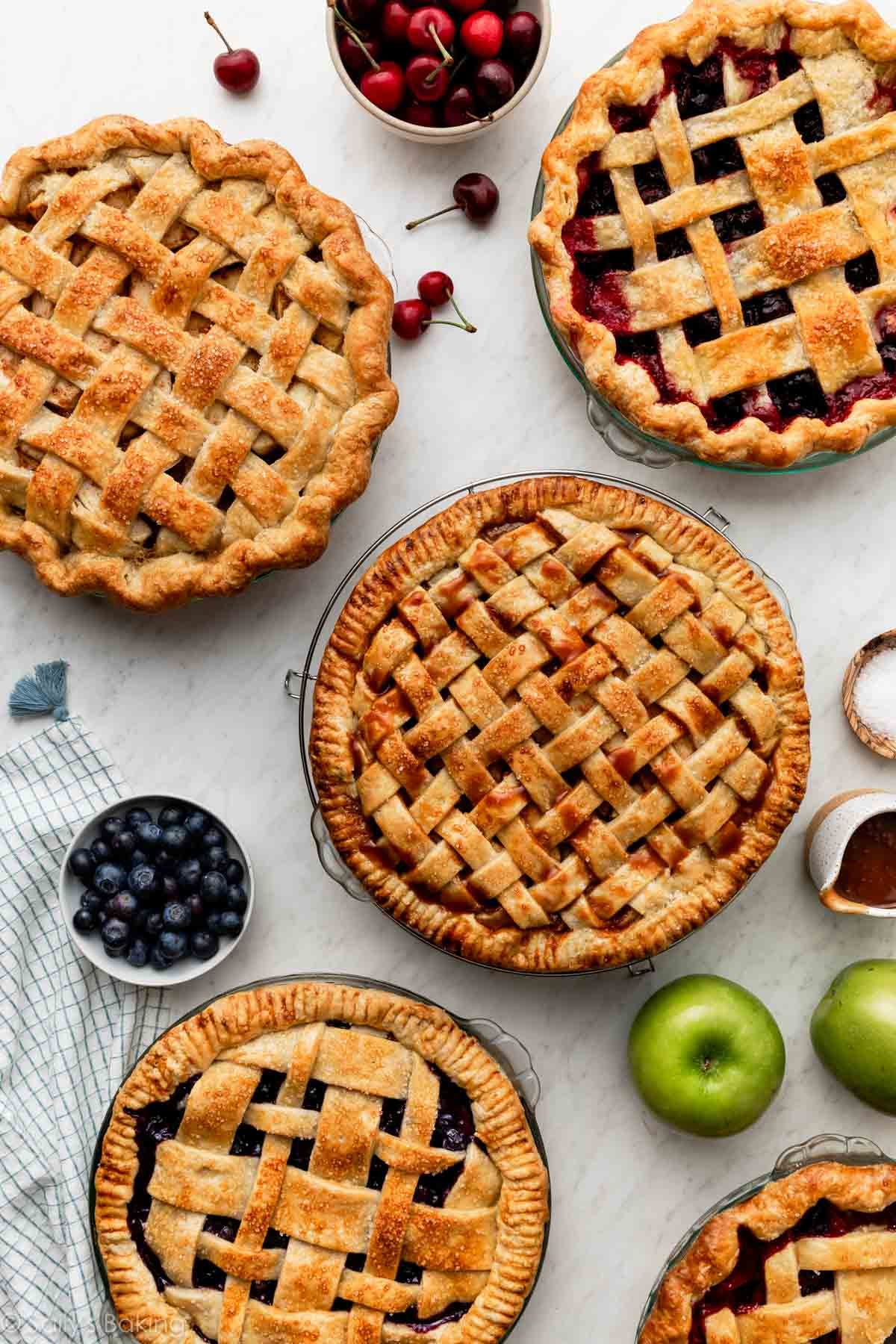 pies with lattice pie crust on marble surface including blueberry pie, 2 apple pies, and cherry pie.