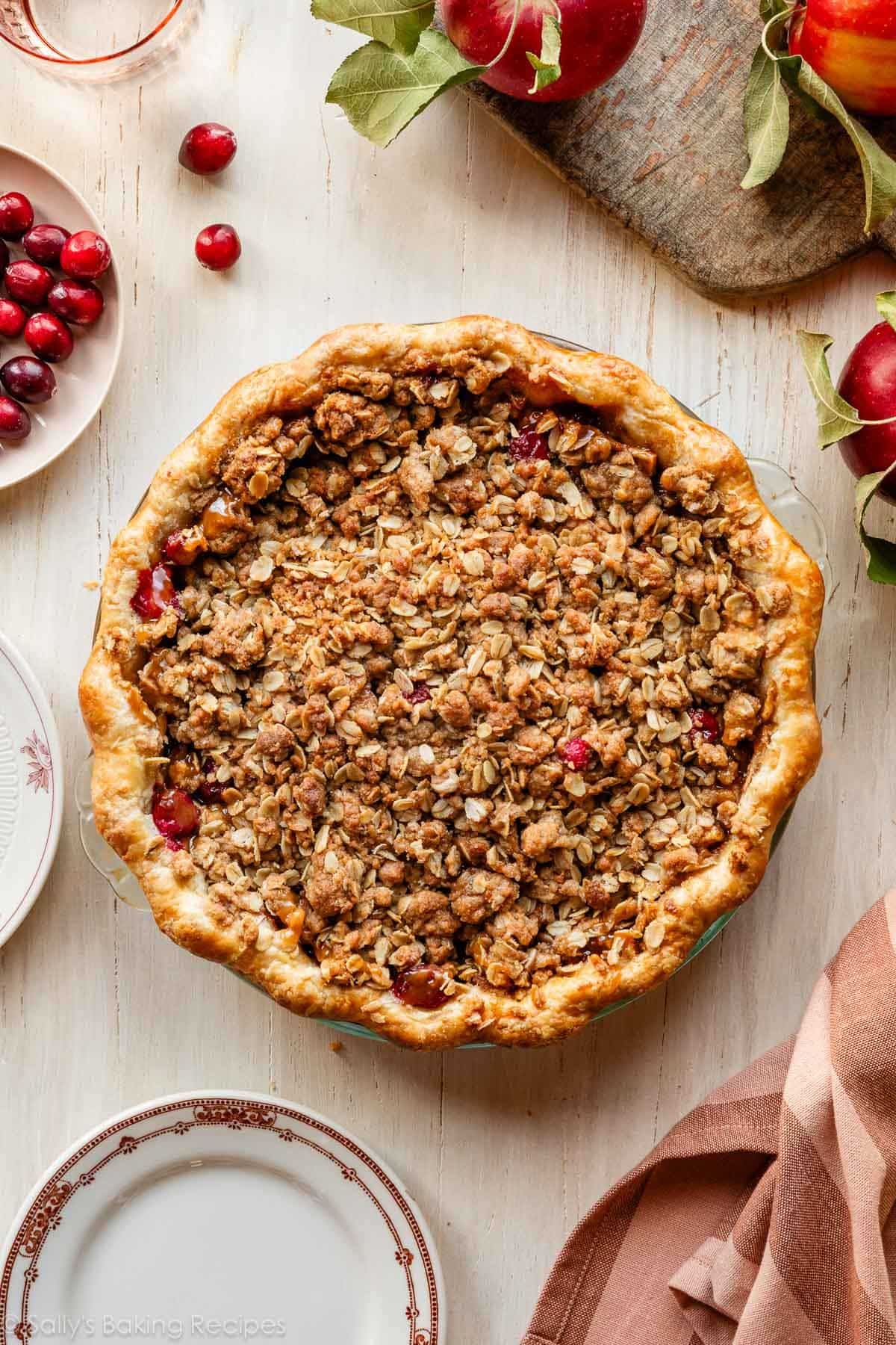 apple cranberry pie with oat crumble topping and apples on wooden surface.