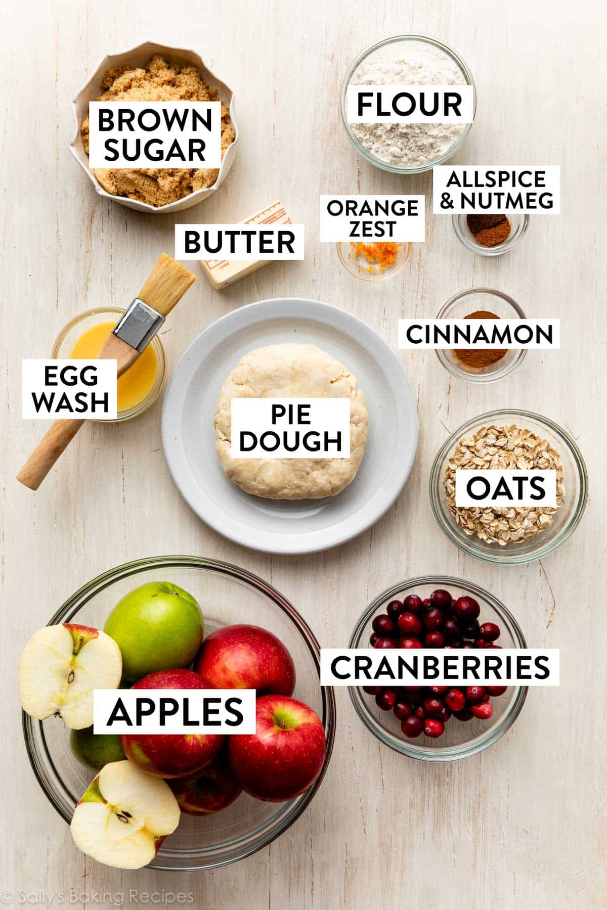 ingredients on white wooden surface including pie dough, oats, brown sugar, eegg wash, apples, cinnamon, and flour.
