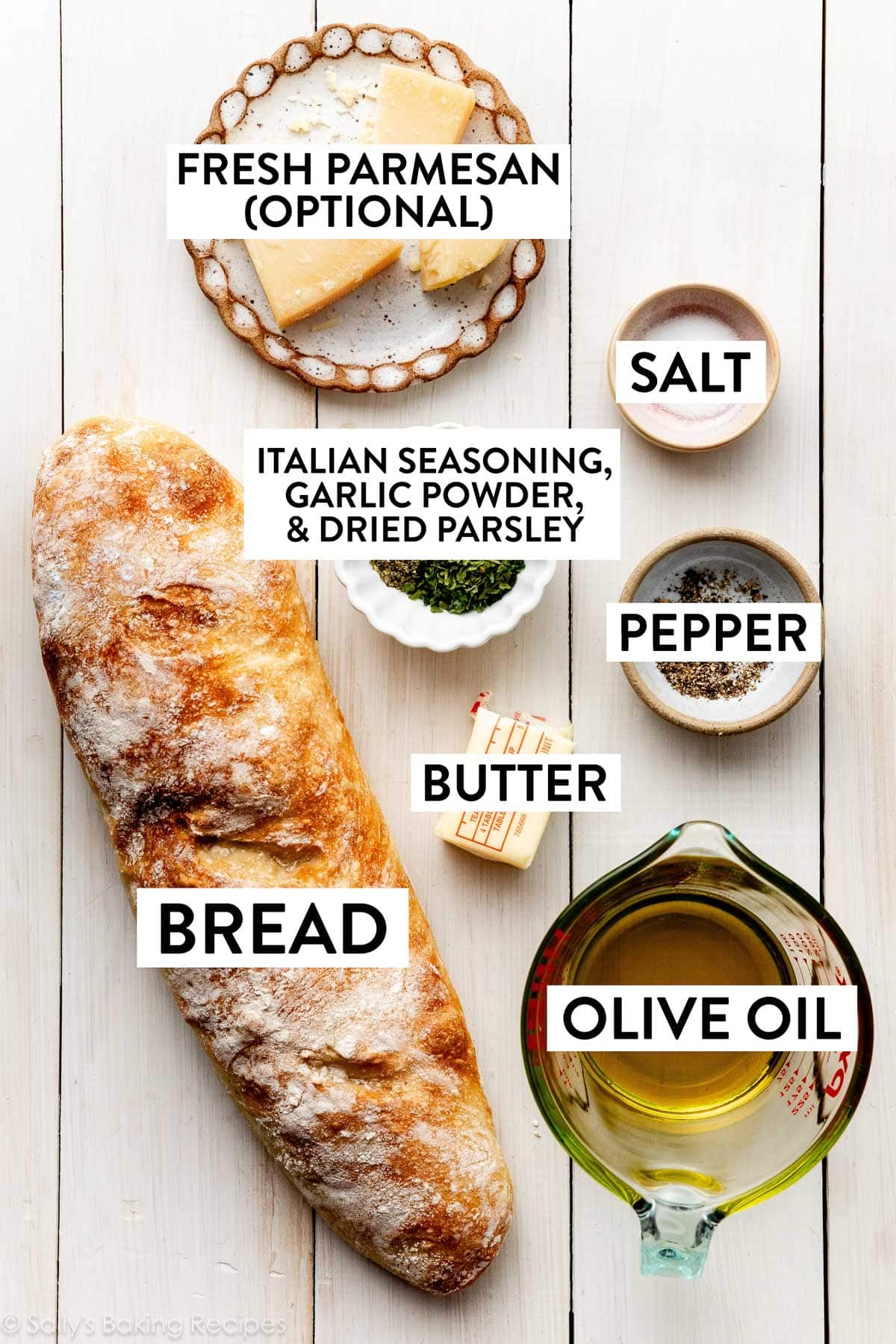 ingredients on white wooden surface including bread, pepper, salt, dried herbs, butter, olive oil, and parmesan cheese.
