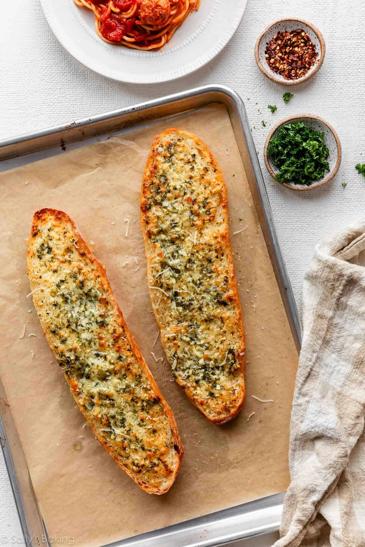baked homemade garlic bread on parchment paper-lined baking sheet with parsley, red pepper flakes, and spaghetti around it.