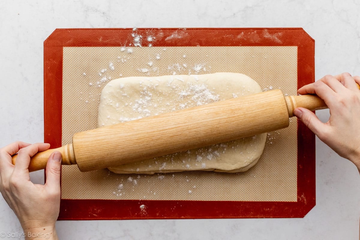 hands rolling folded dough with rolling pin on silicone baking mat.