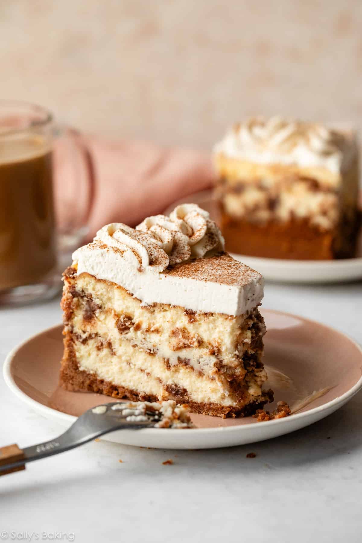 Slice of cinnamon roll cheesecake with whipped cream on top pictured on tan plate.