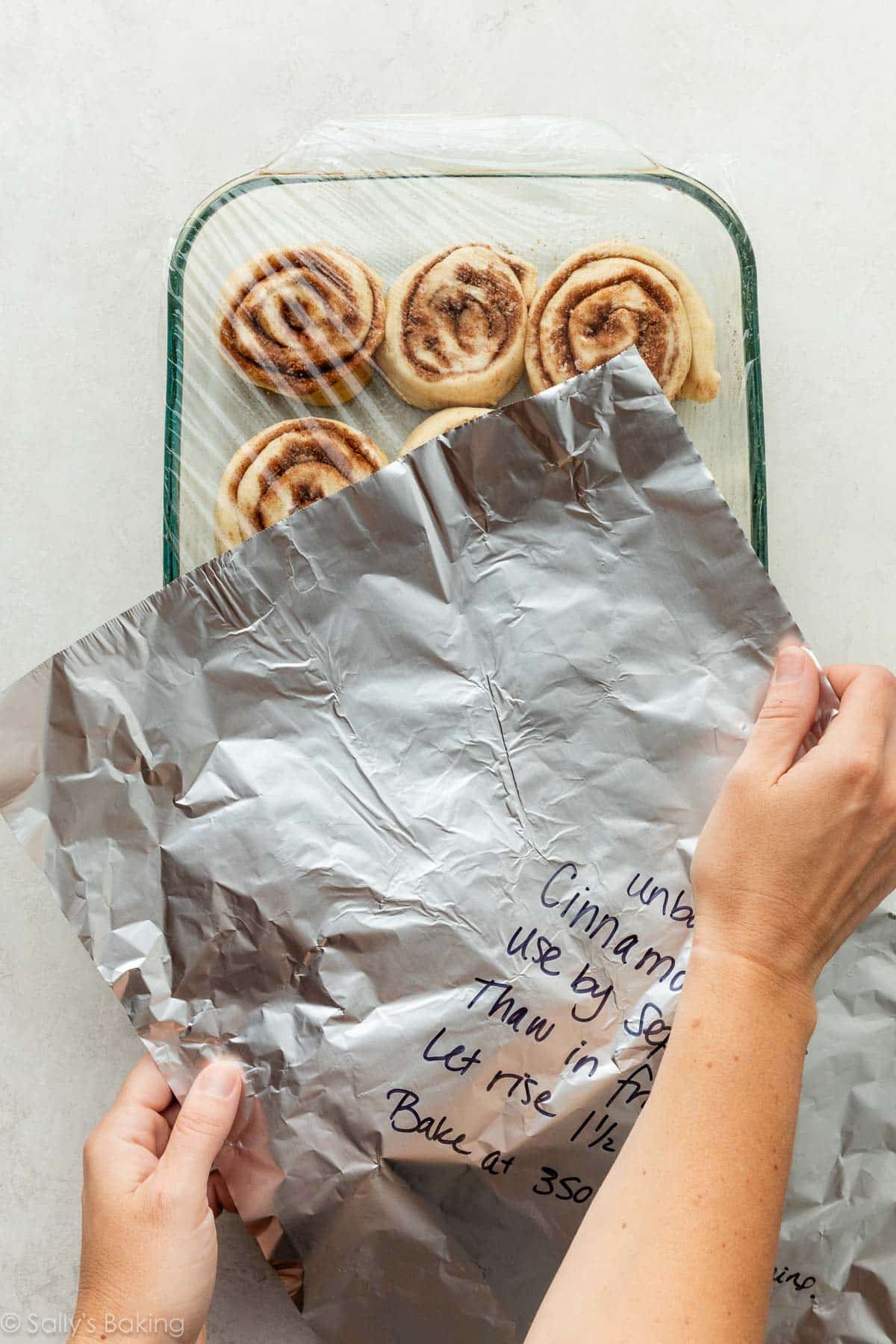 hands covering pan of cinnamon rolls with aluminum foil.