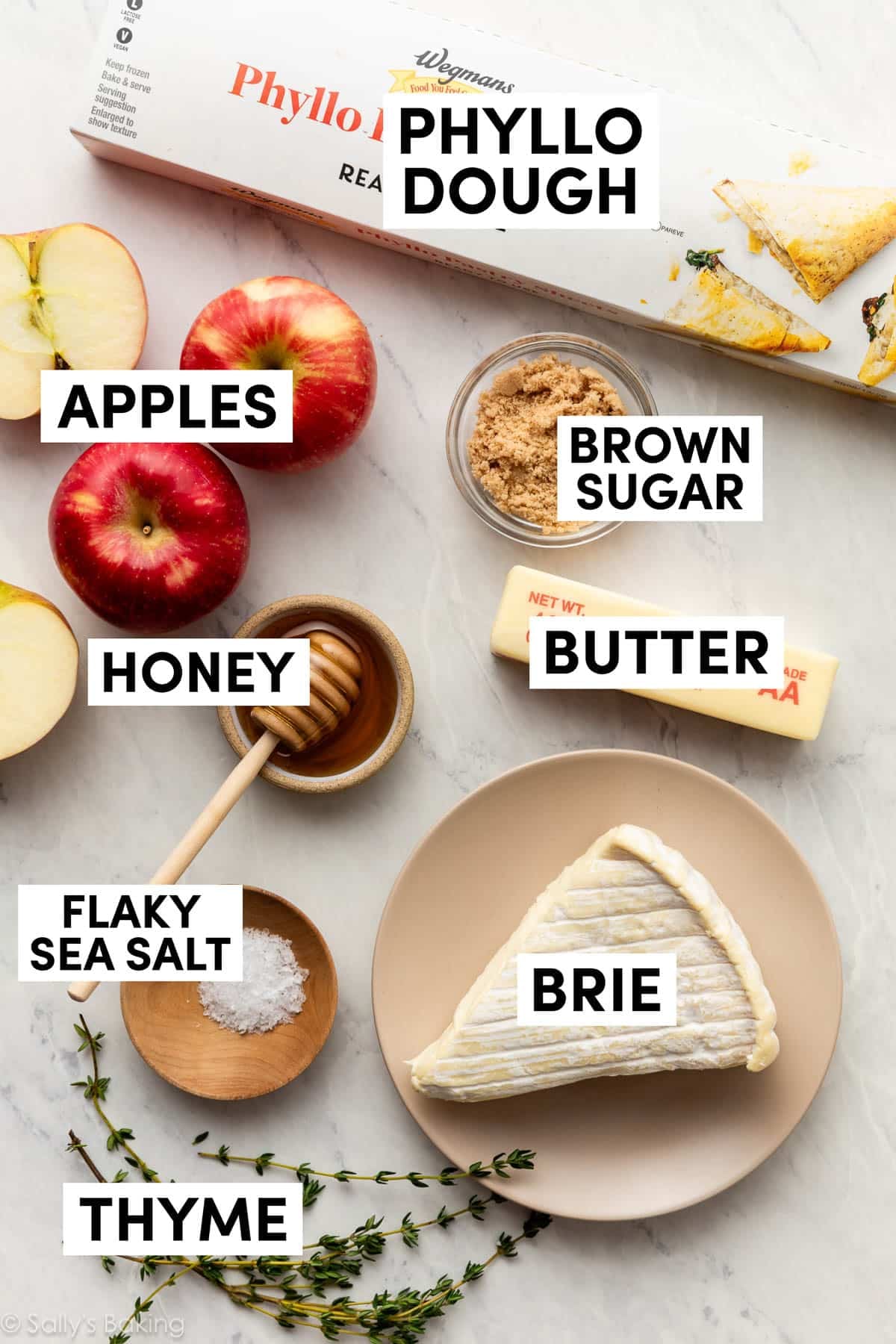ingredients on marble surface including phyllo dough in box, apples, brie, honey, and butter.